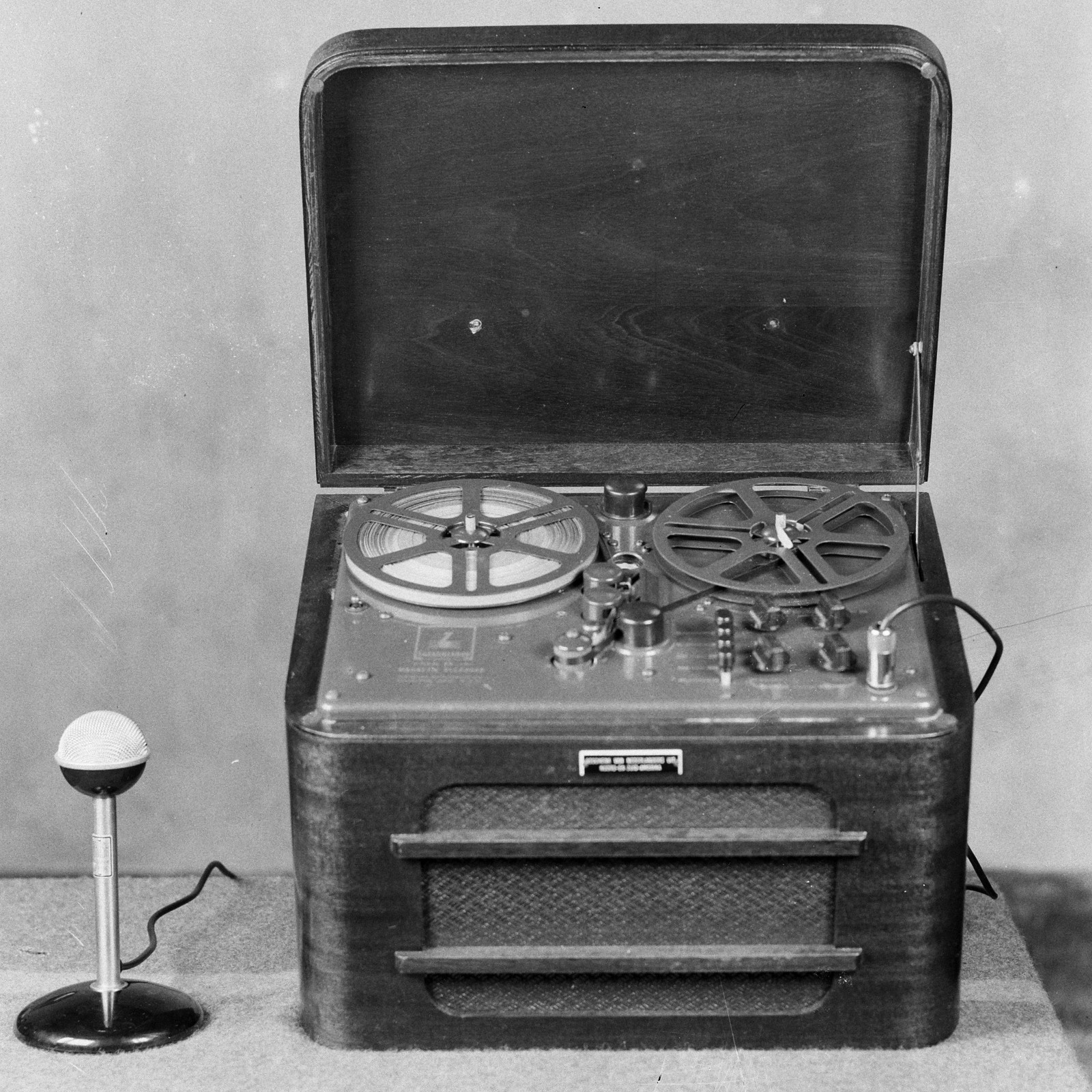 Open portable magnetic-tape recorder with supply and take-up reels, a visible tape path, transport controls, speaker cabinet, and separate microphone.