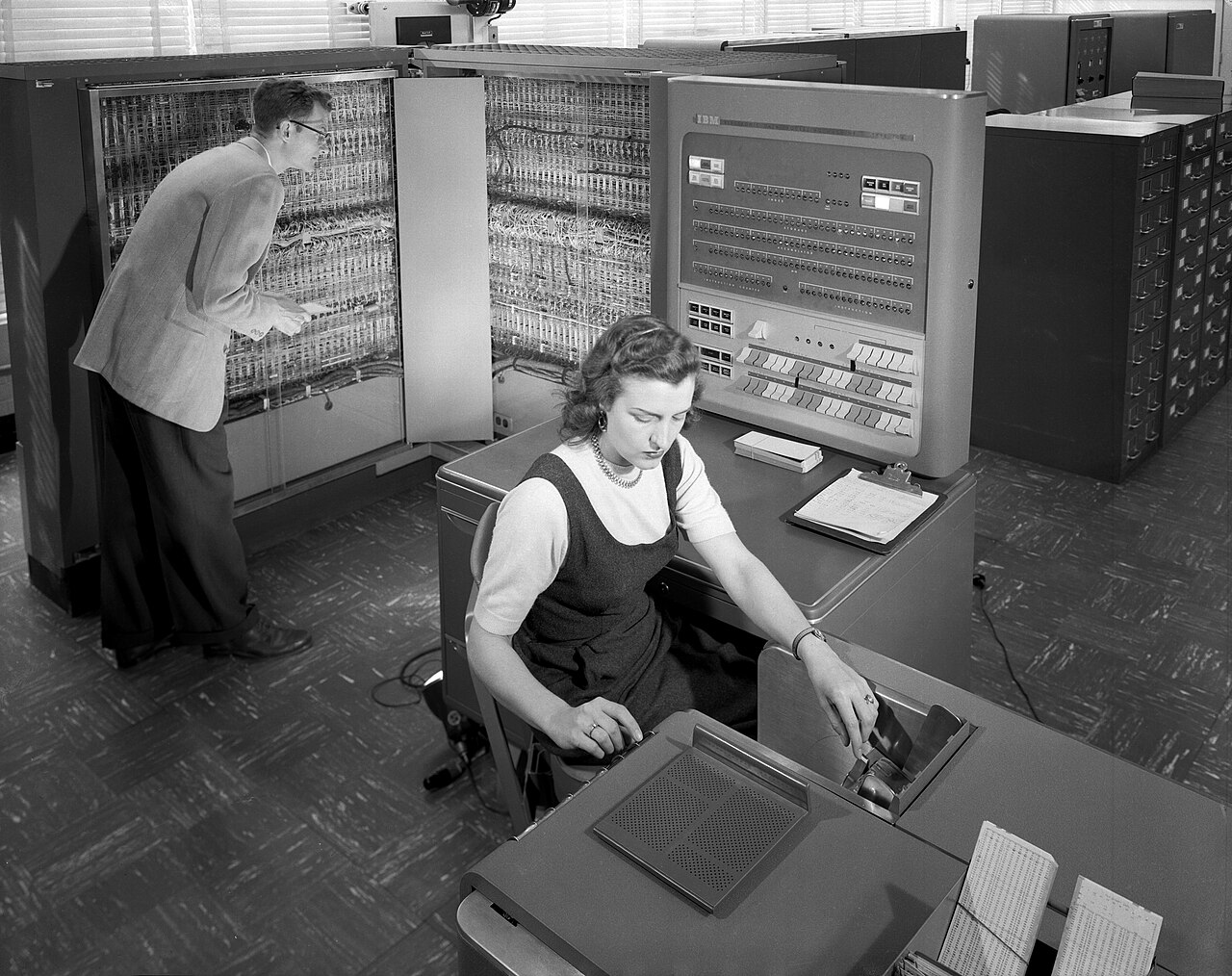 Two operators stand at the control console of a room-sized IBM 704 computer installation.